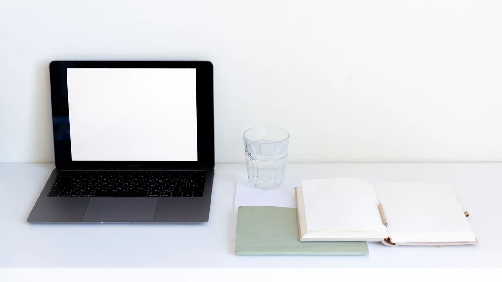 An open laptop with a blank screen on a white desk, beside an empty notebook, a pen, and a glass of water.